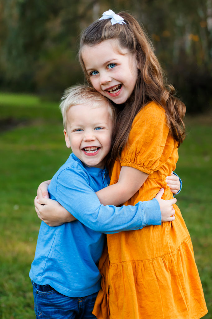 a girl in a yellow dress and a smaller boy in a blue shirt hugging each other with genuine smiles on their faces
